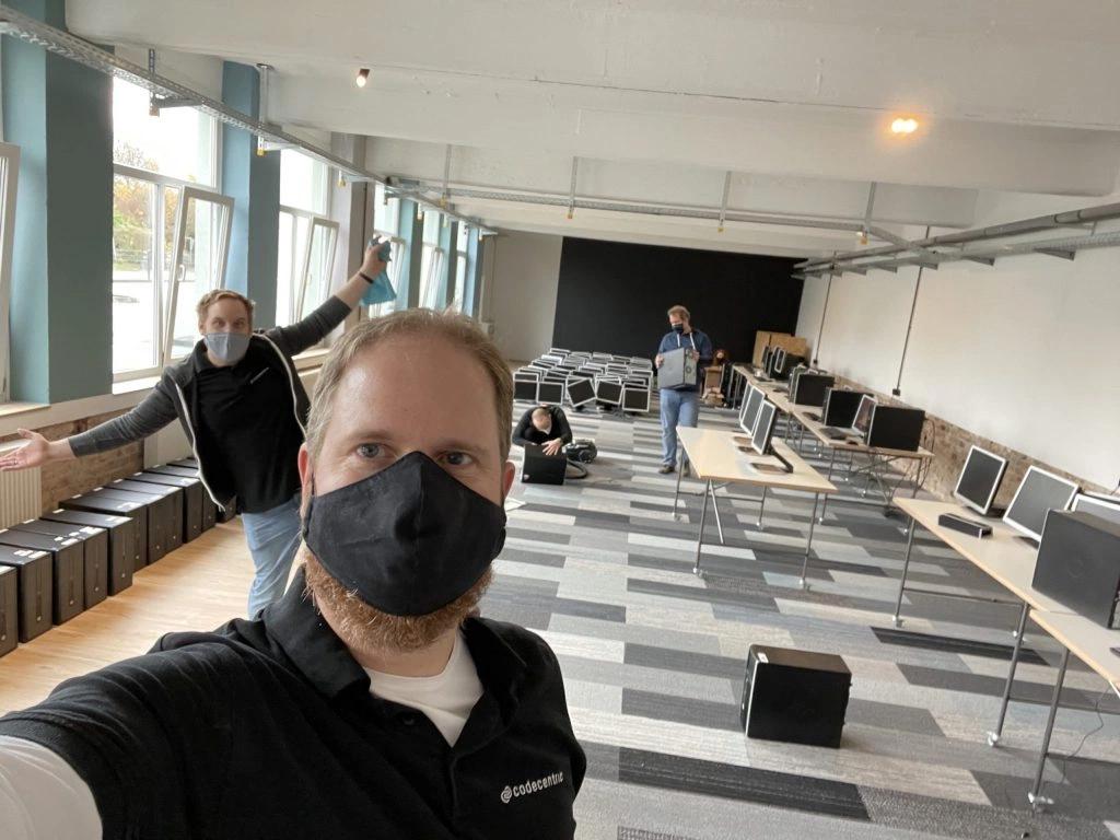 Selfie einer Person mit Maske, im Hintergrund 2 weitere Personen die an Computern arbeiten, eine weitere Posiert fürs Bild.