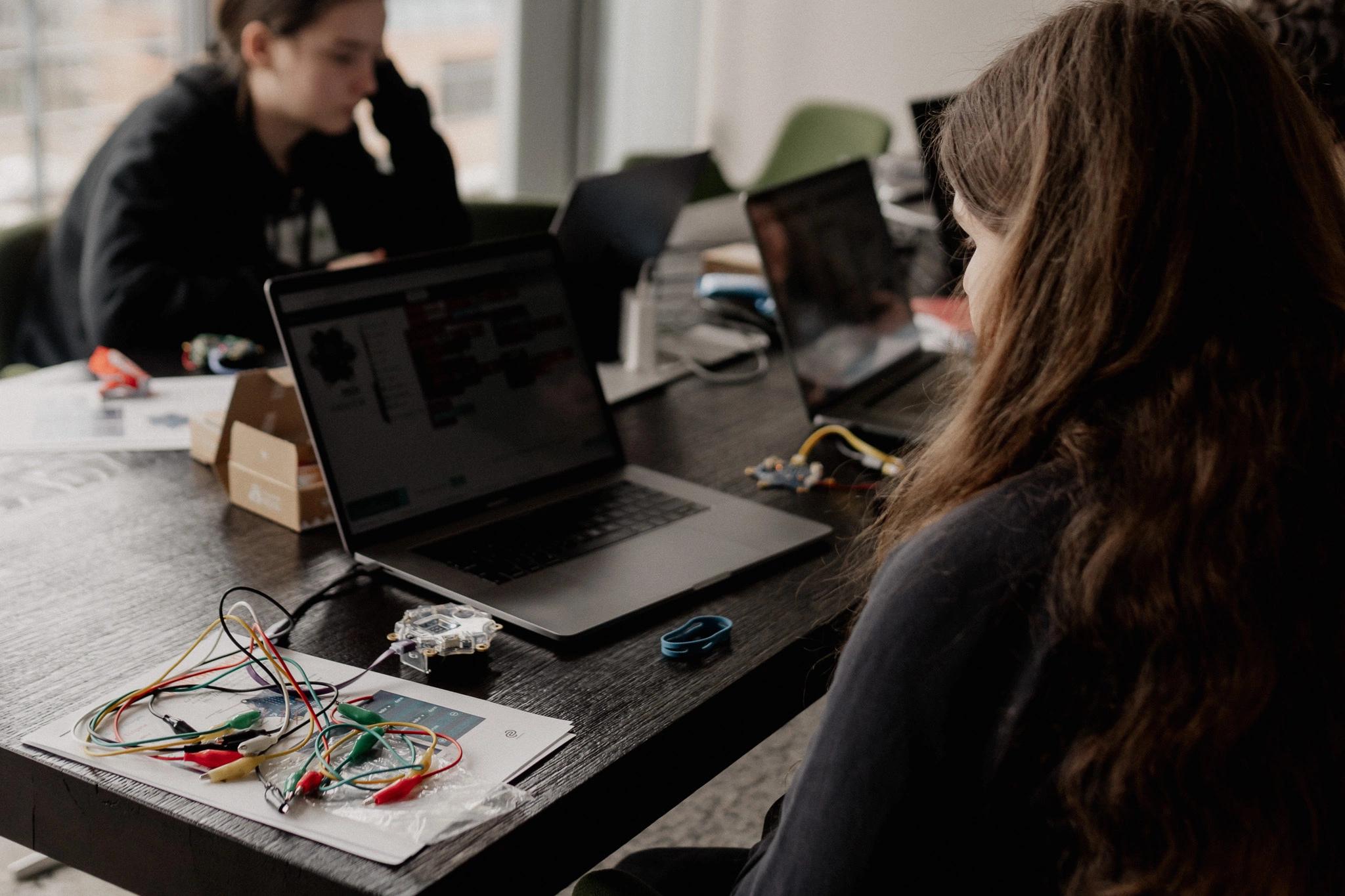 Person arbeitet an einem Laptop an den viele Kabel angeschlossen sind, im Hintergrund weitere Personen an Laptops.