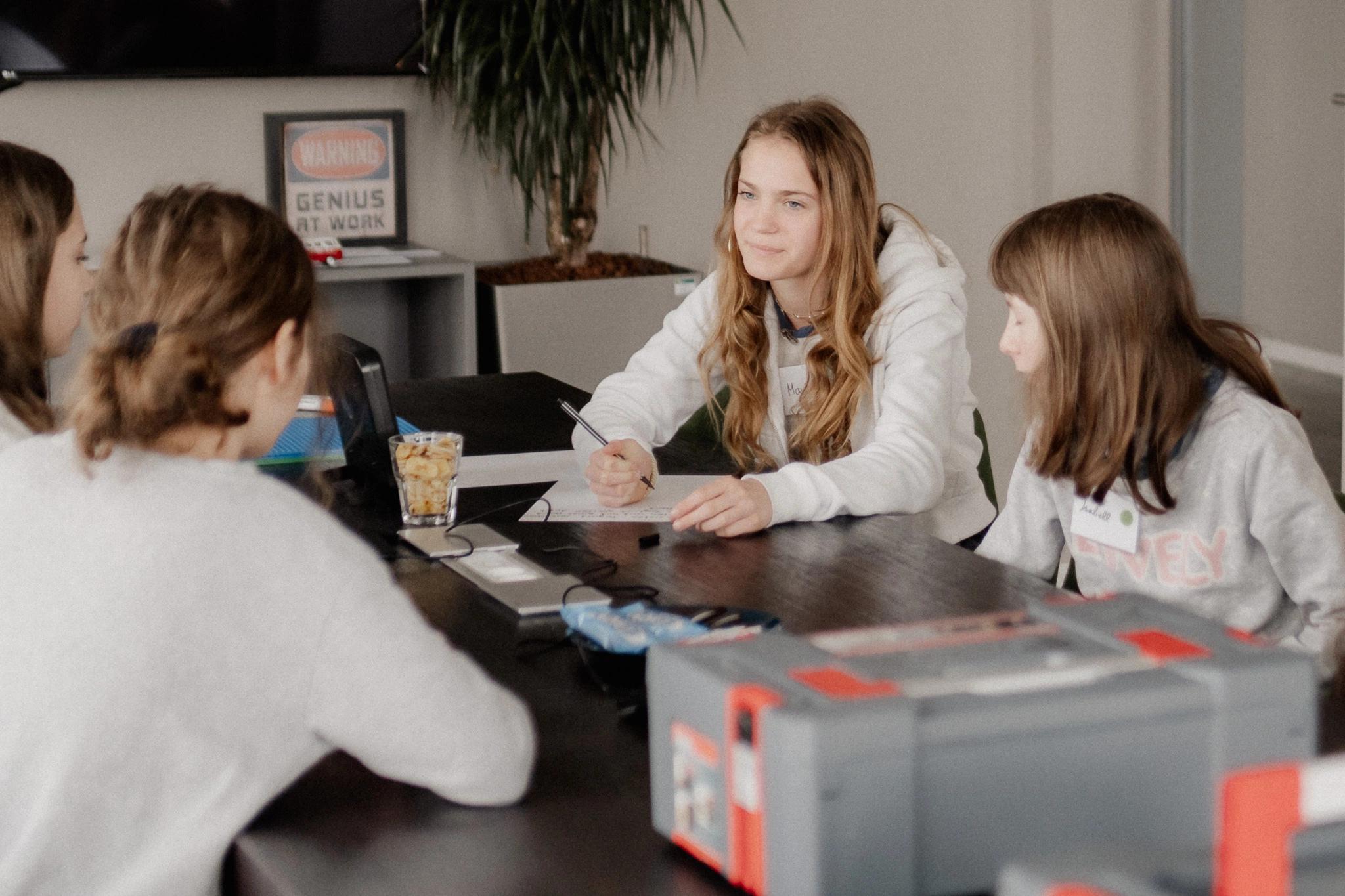 4 Personen an einem Tisch besprechen etwas, eine von ihnen schreibt etwas auf ein Blatt.