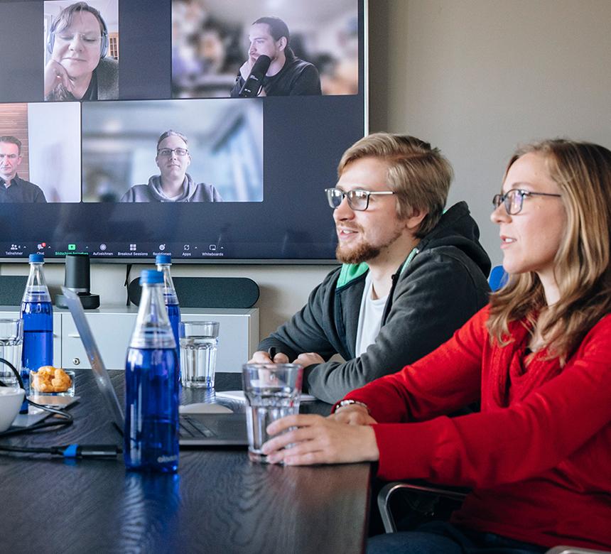 Projektbesprechung: drei Personen in einem Meetingraum, 5 weitere per Videokonferenz auf dem Bildschirm an der Wand dabei