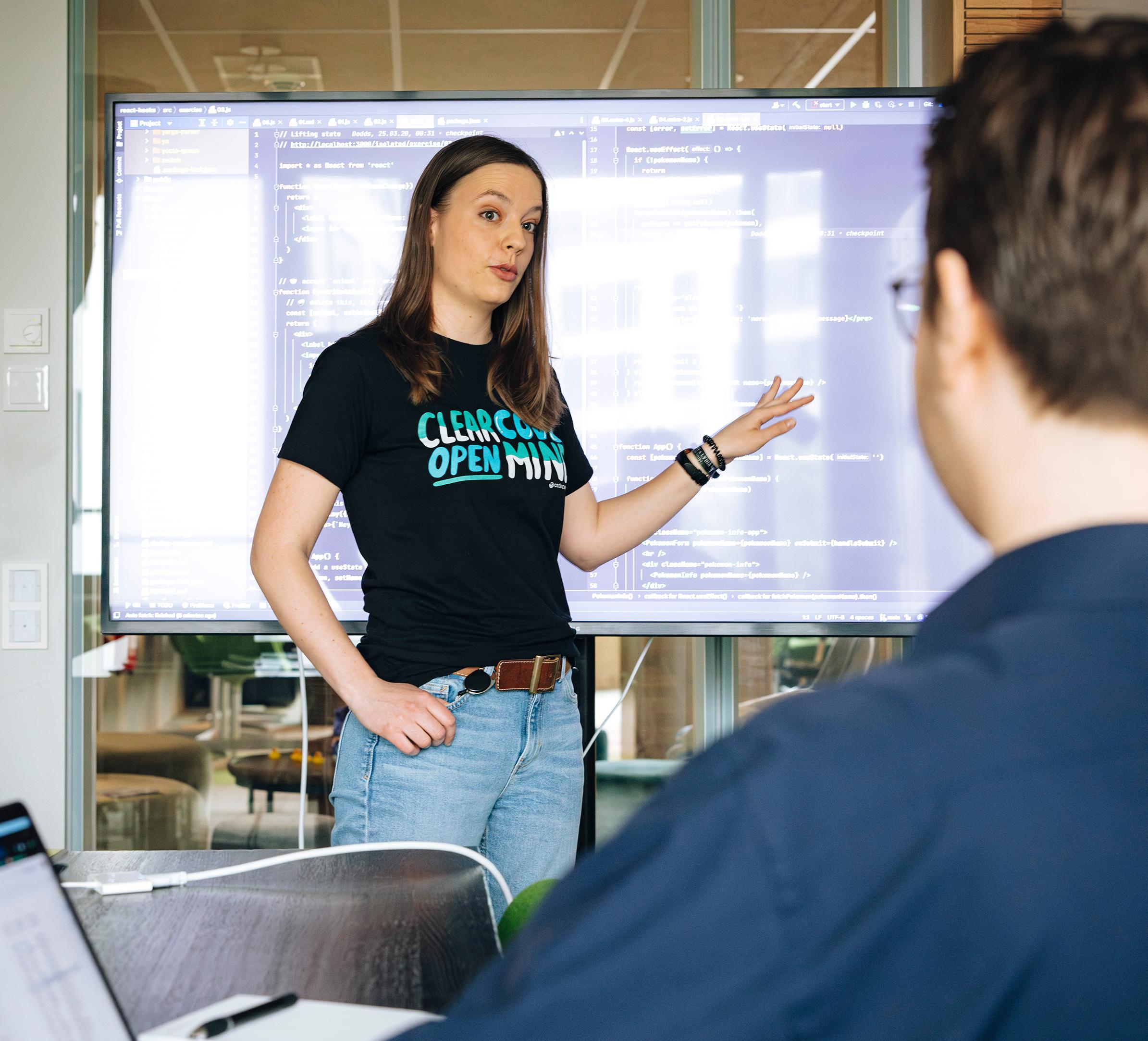 Frau steht vor einem großen Monitor mit Code und erklärt etwas einem Mann, der im Vordergrund von hinten zu sehen ist.