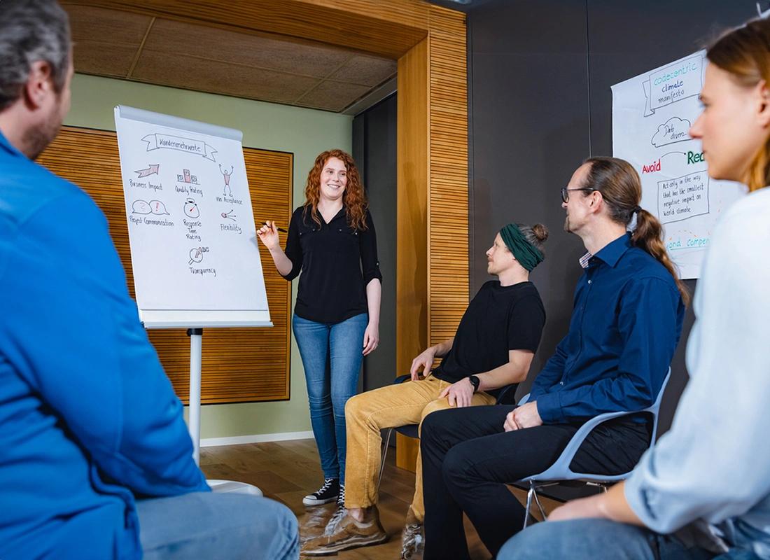 Eine Mitarbeiterin zeigt einer Gruppe von Leuten im Stuhlkreis etwas am Flipchart