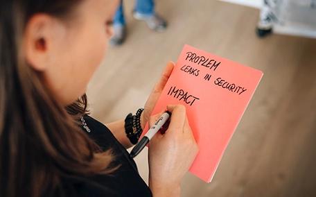 Frau schreibt mit schwarzem Stift auf roten Zettel die Worte "Problem leaks in Security, Impact"