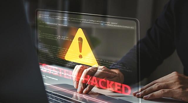 Foto von Monitor mit großem gelben Achtung Schild "System hacked", dahinter Hände auf der Tastatur