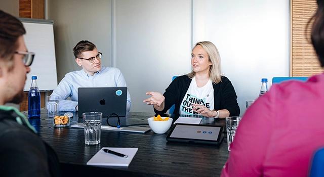 Drei Personen sitzen am Tisch in einem Meetingraum mit Laptop und besprechen sich.