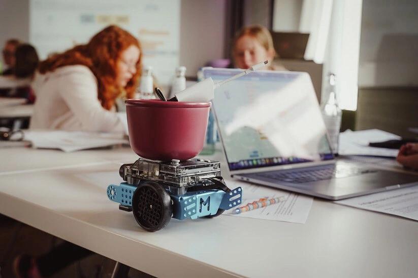 Ein kleiner Roboter auf einem Tisch neben einem Laptop, im Hintergrund 2 Personen unscharf zu erkennen.