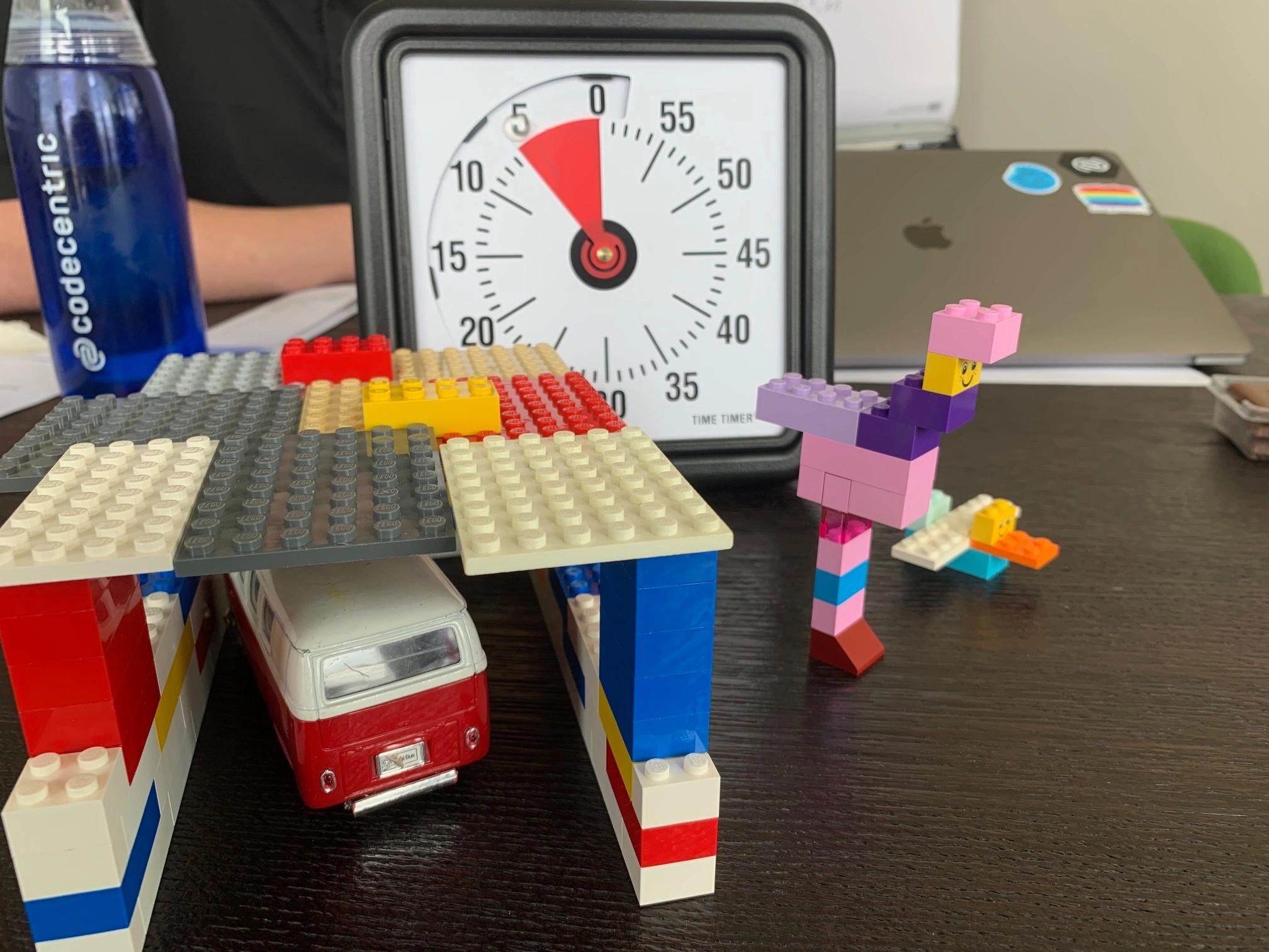 Eine aus Lego gebaute Garage mit einem Spielzeugauto auf einem Tisch, dahinter ein Wecker und eine codecentric Flasche.
