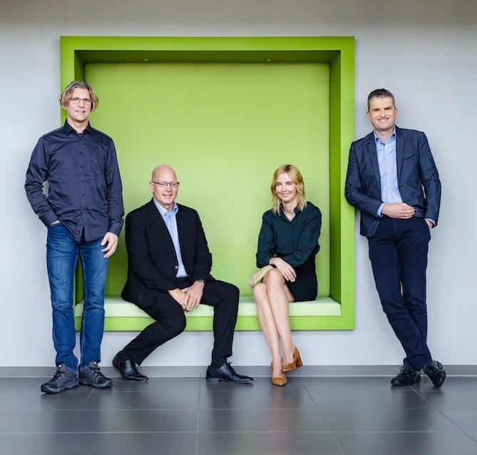 4 Vorstände: Stefan und Verena sitzen in einer in die Wand eingelassenen Box, Lars und Rainer stehen rechts und links davon.